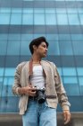 From below young latin man photographer holding photo camera looking away against modern building — Stock Photo