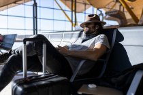 El tipo con sombrero en el aeropuerto en la sala de espera sentado esperando su vuelo, con auriculares inalámbricos para escuchar música, duerme y con su sombrero cubre sus ojos - foto de stock