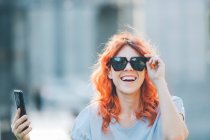 Модна жінка з імбирним волоссям і в сонцезахисних окулярах робить самостріл на мобільний телефон в сонячний день на вулиці — стокове фото