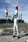 Full body man in spacesuit standing on rocky ground against metal fence and striped rocket shaped antennas on sunny day — Stock Photo