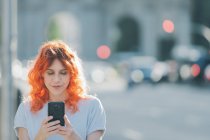 Alegre pelirroja mujer en la calle y mensajería en las redes sociales en el teléfono móvil - foto de stock