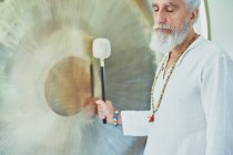 Vista lateral da colheita macho adulto em roupas brancas com martelo jogando gong suspenso durante a prática espiritual — Fotografia de Stock