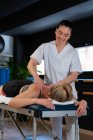 Дружелюбная массажистка, улыбающаяся и массирующая плечи женщины во время работы в клинике физиотерапии — стоковое фото