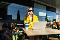 Улыбающаяся азиатская деловая женщина в желтом пальто сидит за столом и пьет кофе со своим смартфоном и ноутбуком — стоковое фото