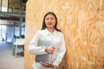 Sorrindo etnia asiática empresária em pé com computador perto da parede no espaço de co-trabalho enquanto olha para a câmera — Fotografia de Stock