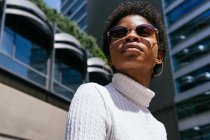 De baixo positivo jovem afro-americano mulher na roupa da moda olhando para longe e desfrutando da luz solar enquanto sentado no banco na rua da cidade moderna — Fotografia de Stock