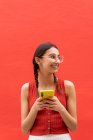 Fröhliche junge Frau mit Zöpfchenfrisur surft auf dem Smartphone und schaut auf rotem Hintergrund auf der Straße weg — Stockfoto