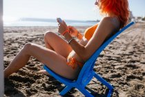 Вид сбоку на анонимную молодую рыжеволосую женщину, использующую телефон, сидя на стуле на пляже в солнечный летний день — стоковое фото