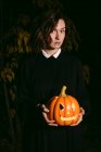 Frau in schwarzem Kleid steht an Halloween mit leuchtender Kürbislaterne im dunklen Wald und blickt in die Kamera — Stockfoto