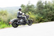 Full body of bearded ethnic male biker in black leather jacket and helmet riding modern motorbike on asphalt road amidst lush green trees growing in mountainous valley — Stock Photo
