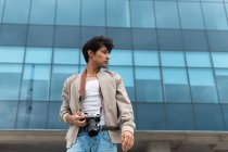 De baixo jovem fotógrafo latino segurando câmera de fotos olhando para longe contra o edifício moderno — Fotografia de Stock