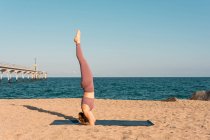 Vista laterale del rilassato bilanciamento femminile a Salamba Sirsasana sul tappeto mentre fai yoga sulla riva del mare — Foto stock