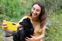 Mulher alegre tomando auto tiro com cachorro fofo preto ao usar o smartphone e sentado em banco de madeira no campo — Fotografia de Stock
