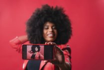 Улыбающаяся афроамериканка с африканской прической, фотографирующая себя на мобильном телефоне на красном фоне в студии — стоковое фото