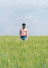 Hombre tranquilo con torso muscular desnudo tocando la parte superior de la hierba caminando en el campo verde contra el cielo nublado - foto de stock