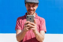 Content young bearded hipster guy in casual polo shirt and cap browsing mobile phone while standing against blue wall in sunlight — Stock Photo