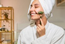 Giovane donna felice con asciugamano sulla testa sorridente e massaggiante viso con rullo di giada durante la routine di cura della pelle a casa — Foto stock