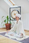 Mulher madura com olhos fechados sentados com pernas cruzadas no tapete fofo enquanto pratica ioga em casa — Fotografia de Stock