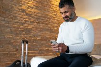 Низький кут позитивного врожаю етнічного чоловіка, який сидить на ліжку біля валізи та переглядає мобільний телефон у готельному номері — стокове фото