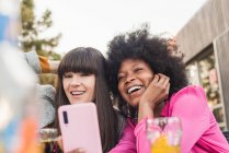 Багаторасовий пара гомосексуальних жінок, які роблять самостріл на мобільний телефон, сидячи за столом з коктейлем у вуличному кафе — стокове фото