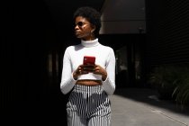 Afroamerikanerin in trendiger Kleidung surft auf ihrem Handy in den sozialen Medien, während sie an sonnigen Tagen auf der Straße der Stadt spaziert — Stockfoto