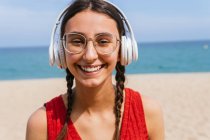Portrait of happy female in headphones listening to music on sandy seashore on sunny day in summer looking at camera — Stock Photo