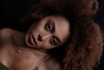 Modelo feminino afro-americano encantador com cabelo encaracolado olhando para a câmera no estúdio escuro — Fotografia de Stock