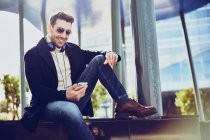 Conteúdo masculino em vestuário elegante com fones de ouvido e celular sentado na cidade no dia ensolarado — Fotografia de Stock