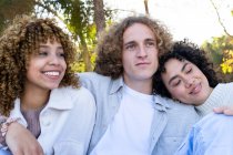 Bajo ángulo de hombre abrazando a diversas amigas que tienen el pelo rizado mientras se relajan en el parque mirando hacia otro lado - foto de stock