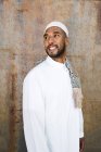 Hombre islámico en ropa blanca auténtica mientras está de pie contra la pared grumosa - foto de stock