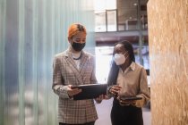 Multiethnic female colleagues with tablets and in protective masks standing in coworking space and discussing project — Stock Photo