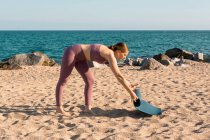 Vue latérale complète du corps de la jeune femme en vêtements de sport plaçant tapis de yoga sur le sable tout en se préparant pour la pratique sur la plage près de l'océan — Photo de stock