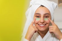 Mujer feliz con toalla en la cabeza sonriendo y extendiendo máscara verde en la cara mientras mira el espejo en el baño en casa - foto de stock