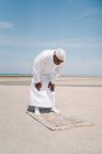 Niño completo varón islámico en ropa blanca tradicional de pie sobre una alfombra y rezando contra el cielo azul en la playa - foto de stock