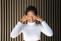 Mulher afro-americana feliz em camisola na moda cobrindo os olhos com sorriso contra a parede listrada na rua — Fotografia de Stock