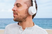 Unbekümmerter junger bärtiger Typ in stylischem lässigem Hemd, der über drahtlose Kopfhörer Musik hört und frische Brise genießt, während er den Sommertag am Sandstrand am Meer verbringt — Stockfoto