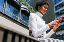 Bajo ángulo de joven hembra negra con ropa elegante sentada en el banco y navegando por el teléfono celular en un día soleado en la calle moderna de la ciudad - foto de stock