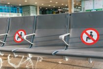 Fila de asientos vacíos con marcadores de restricción roja para el distanciamiento social en la sala de salidas del aeropuerto durante la epidemia de COVID - foto de stock