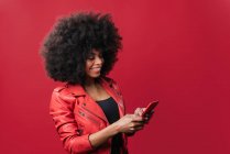 Захоплена афроамериканка з африканською зачіскою переглядає мобільний телефон на червоному фоні у студії — стокове фото