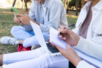 Ritagliato uomo e donne multietniche irriconoscibili seduti sul prato nel parco utilizzando il computer portatile e il blocco note di condivisione — Foto stock