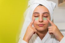Donna felice con asciugamano sulla testa sorridente e diffusione maschera verde sul viso mentre si guarda specchio in bagno a casa — Foto stock
