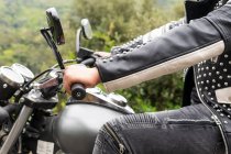 Crop mâle motard en veste et casque en cuir noir à moto moderne sur route asphaltée au milieu d'arbres verts luxuriants poussant dans la vallée montagneuse — Photo de stock