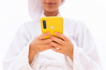 Crop Young female in bathrobe and towel smiling and browsing mobile phone while resting on balcony after shower — Stock Photo