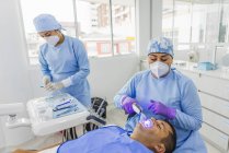 Высокий угол сфокусированного врача в медицинской форме лечащего клиента с помощью стоматологического инструмента с ассистентом, готовящим инструменты в больнице — стоковое фото