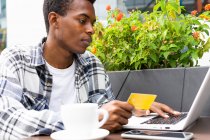 Focado afro-americano masculino pagando por ordem com cartão de plástico ao usar laptop durante compras on-line no café de rua — Fotografia de Stock