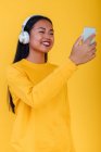 Contenido Mujer asiática escuchando música en auriculares y tomando fotos en el teléfono inteligente sobre fondo amarillo en el estudio - foto de stock