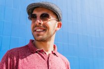 Young bearded guy wearing casual red polo shirt and cap looking at camera over sunglasses and smiling friendly while standing against blue wall on street in summer day — Stock Photo