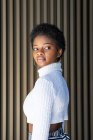 Неемоційна афро-американська жінка в модних светрах, дивлячись на смугастий будинок стіни на вулиці — стокове фото
