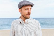 Young bearded male in trendy casual clothes and cap standing on sandy beach towards waving sea while spending summer holidays on seashore — Stock Photo