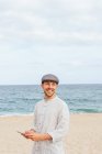 Позитивний молодий бородатий хлопець у стильній сорочці і кепці посміхається і дивиться геть, переглядаючи мобільний телефон на піщаному пляжі біля моря — стокове фото